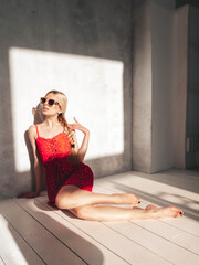 Young beautiful smiling female in trendy summer red dress. Sexy carefree blond woman sitting near wall in studio. Cheerful and happy in sunglasses. At sunny day. Shadow from window
