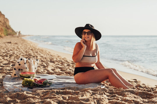 Smiling Young Woman Looking Away and Talking on the Phone While Sitting on Sandy Beach by the Sea