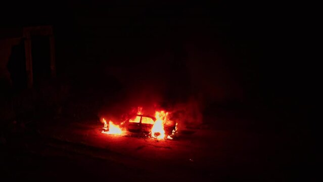 Drone aerial shot of burning car. Exploded destroyed parked old car on fire. Horror or thriller movie scene. Cinematic. Night lights.