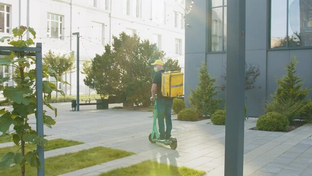 A delivery man with a yellow backpack drives a e-moped into the video frame, stops amd looking around, the man is shot from the back. Excellent delivery during quarantine