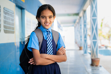 Happy smiling girl kid in school uniform confidently standing at corridor with arms crossed by looking at camera - concept of education, knowledge and childhood development