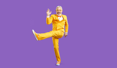 Happy funny energetic stylish senior man having fun in studio. Cheerful mature guy wearing bright yellow party suit with bow tie enjoying music and dancing isolated on solid purple background