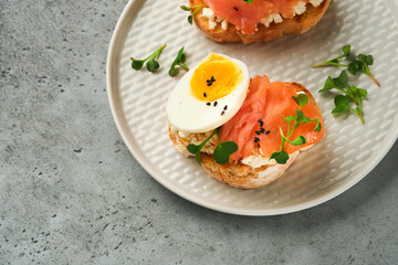 Sandwich with delicious toppings smoked salmon, eggs, herbs and microgreens radish, black sesame seeds over white plate on gray concrete table background. Healthy open sandwich superfood. Top view.