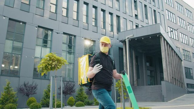 Food supplier with termo backpack riding on electric scooter on the street. A masked courier delivers food in a pandemic period. Delivery man looking at the smartphone screen at the street.