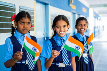 Smiling girl kis in unifrom waving Indian falg by looking at camera at school corridor - concept of...