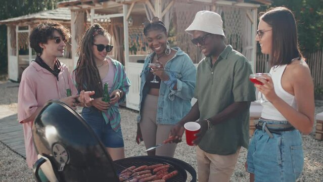 Medium long of five multiethnic young people having BBQ party on beach in summer heat, talking, smiling, roasting sausages on grill