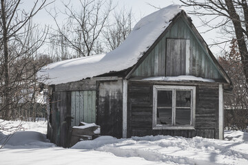 An old abandoned wooden house or cottage with a broken window, covered with snow and impassable snowdrifts. An abandoned house in winter.