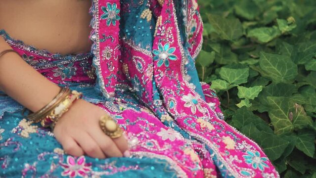Close-up image beautiful young female hands. Hand woman gently touches Indian vintage national pink blue saree dress. Traditional jewelry. Gold bracelets, precious rings on fingers. Backdrop green ivy