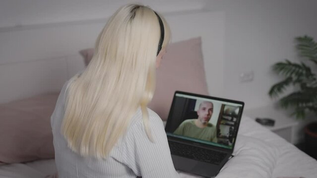A blonde woman sits on a bed and talks on a video call on a laptop with friend.