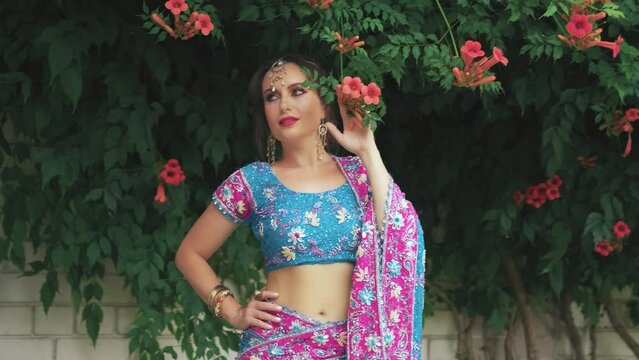 Oriental woman walks towards flowering shrub Kampsis. Indian adult girl princess enjoy scent natural flower. Blue pink traditional vintage dress clothing. Silver tiara. National bracelets adorn hand.