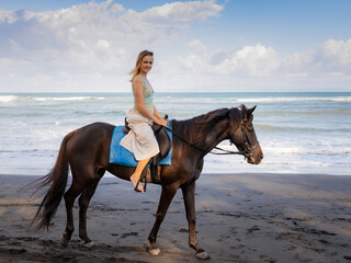 Beautiful woman riding horse on the beach. Outdoor activities. Caucasian woman wearing skirt. Traveling concept. Cloudy sky. Sea view. Copy space. Horizontal layout. Bali, Indonesia