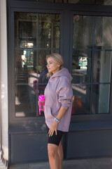 sports activity. blonde girl in violet oversize hoodie and cycling shorts is standing near park cafe background and looking away with smile and pink bottle in hand. sport lifestyle concept, free space