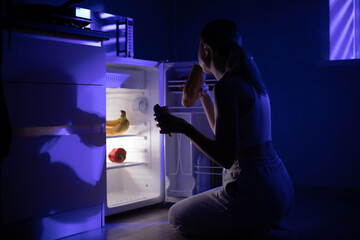A woman eats bread and sausage while sitting near an open refrigerator in the middle of the night.