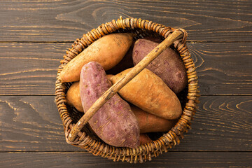 Sweet orange and purple potato tubers in a wicker basket close up on a wooden background, top view
