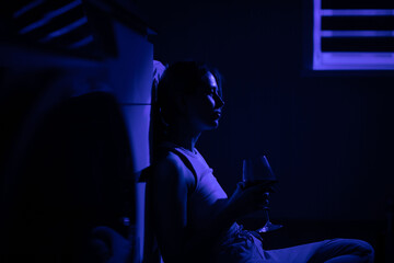 Fototapeta na wymiar Sad woman drinking wine while sitting near the refrigerator in the dark.