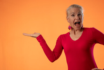 Portrait of beautiful smiling mature woman on orange color background, with copy space, finger hand point to product or sale.