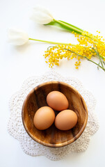 Brown raw chicken eggs in a wooden plate on a lace doily. Easter banner with copy space with tulips and mimosa on white background