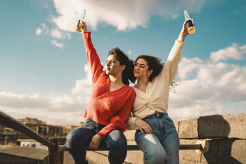 Two happy young women celebrate with beer bottles raised to the sky, sitting on a railing outdoors. Concept of freedom, friendship, youth and celebration.