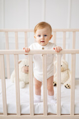 a 6-month-old blond boy stands in a crib in a bright children's room and smiles in a cotton bodysuit, the concept of children's goods