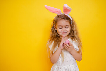 cute smiling blonde girl with bunny ears holding an easter egg in her hands, on a yellow background studio, space for text, kid celebrate easter