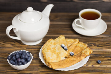 Pancakes are thin homemade pancakes with berries, with a white teapot and a mug of tea on a wooden table. traditional Russian cuisine. Selective focus.
