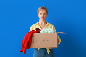 Young woman holding box with donation clothes on blue background