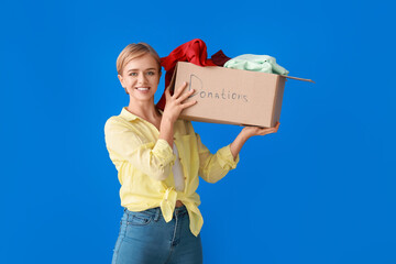 Young woman holding box with clothes for donation on blue background