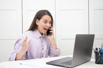 The girl is sitting at the table near the laptop and fooling around, the girl is cheerful, the...