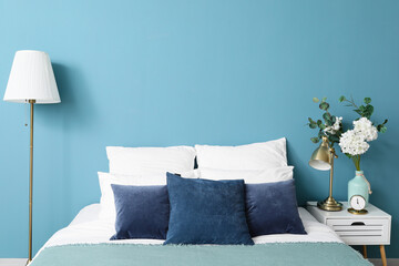 Comfortable bed, vase with flowers on nightstand table and modern lamp near blue wall