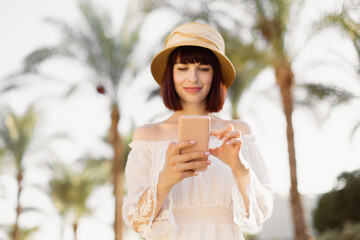 Happy traveler caucasian woman in white dress using mobile phone at tropical city. Summer, holiday, technology and lifestyle concept. Beautiful woman in summer dress at sunny day with phone.