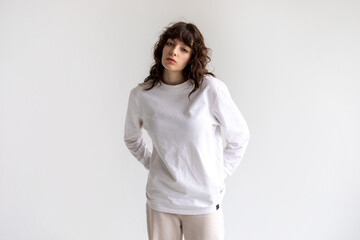 Young woman with curly hair in a white long sleeve t-shirt stands on a white background. Mock-up.