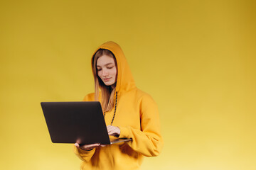 portrait of a beautiful student girl working with a laptop. The girl is dressed in a yellow hoodie. Photo on a yellow background