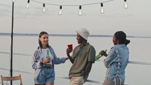 Tilting up of Black guy talking to African American and Biracial girls, friends standing on pier, drinking, having lake party in summer evening