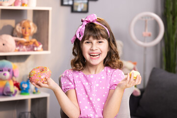 Portrait of child girl choosing between two donuts in home room. Cheerful school girl playing with cakes indoors. Funny teenager girl having fun with colorful donuts at modern home