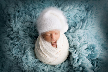 Sleeping newborn baby boy in the first days of life in a white soft cocoon with a knitted woolen white hat on a blue background. Studio macro photography, portrait of a newborn. Woman's happiness.