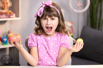 Little girl in pink dress holding donut and macaron in hands. Cute adolescent school girl plays with sweet donut macaroon doing happy fun face expressions on background. Funny concept with sweets.