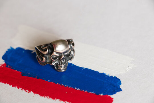A silver ring with a skull lies on a painted Russian flag on white paper, death and war, sanctions and the death of civilians