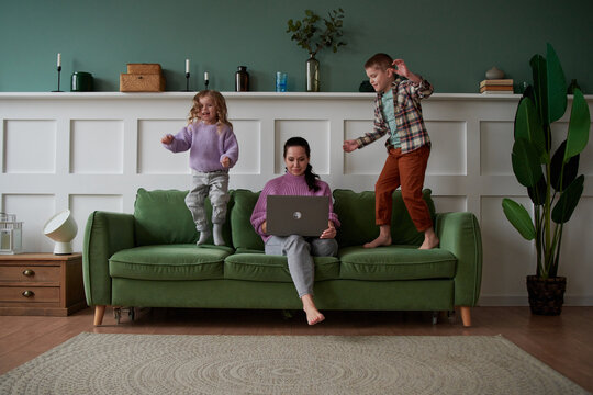 Serious woman using laptop near playing children at home.