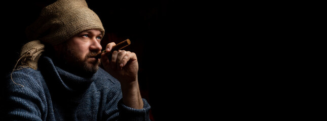Middle-aged man with a cigar in a hat. Brutal unshaven man. Dark background. Banner. Place for text.
