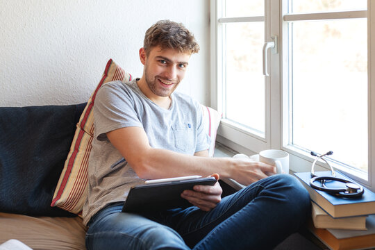 Junger Medizin-Student sitzt zu Hause mit einem digitalen Tablet auf seinem Bett vor einem Fenster und lernt. Er greift gerade zu eine Tasse und lacht in die Kamera. 