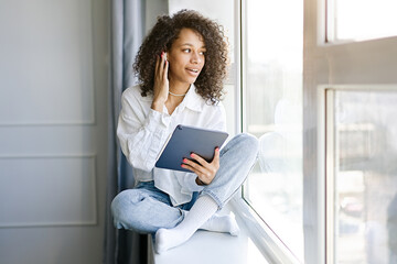 Cute african american girl having a video chat through tablet while sitting at home in the living room by the window