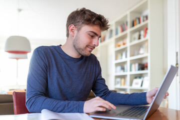 Junger Mann, Student, sitzt zu Hause am Schreibtisch mit einem Laptop und arbeitet online.