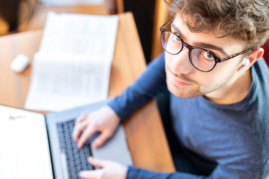 Junger Mann sitzt am Schreibtisch mit seinem Laptop und arbeitet, Blick von oben, er schaut in die Kamera.