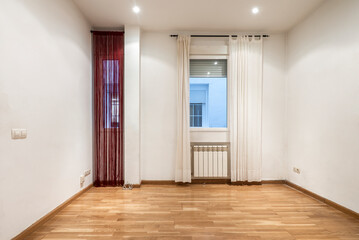 Empty living room with oak parquet flooring, with aluminum windows, radiators and red and white curtains