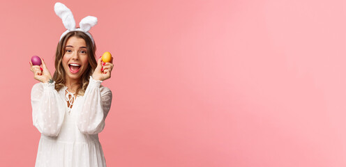 Holidays, spring and party concept. Portrait of excited charming young woman celebrating Easter in rabbit ears and white party dress, dancing with two painted eggs, pink background