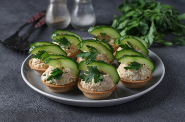 Tartlets with cod liver, egg, garnished with cucumber and parsley on plate on gray background