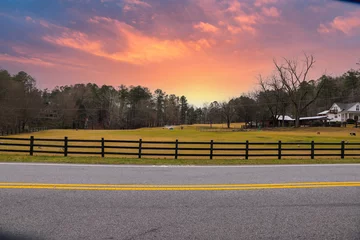  a farm with yellow and green grass surrounded by a street with a yellow line, a black wooden fence and lush green and bare winter trees with animals grazing and a purple and pink sunset in Marietta © Marcus Jones