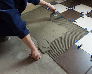 A worker applies adhesive with a notched trowel to lay ceramic tiles on a floor.