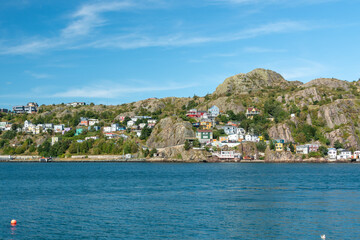Fototapeta premium The hillside of St. John's Harbour, on a sunny day, under blue sky and white clouds. The colorful wooden houses are scattered along the hillside with the blue ocean in the foreground. 