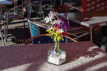 An empty clear glass tequila bottle on an outside restaurant patio table with chairs and shutter glass doors. The bottle is being used as a vase holding colorful red, white and yellow flowers.
